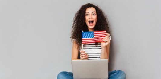 Idiomas e Carreira: Por que aprender inglês pode abrir portas mulher sentada no chão com notebook no colo mostrando bandeira dos Estados Unidos